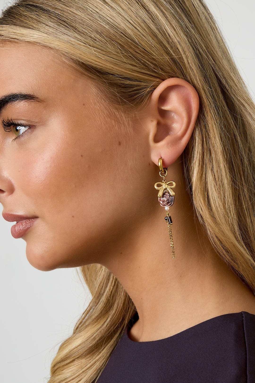 Close-up side view of blonde woman wearing gold-tone floral drop earring with bow, purple flower bead, faux pearls, and chain detail