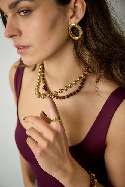 Close-up of woman wearing layered bead necklace and matching gold hoop earrings and ring, dressed in dark burgundy top