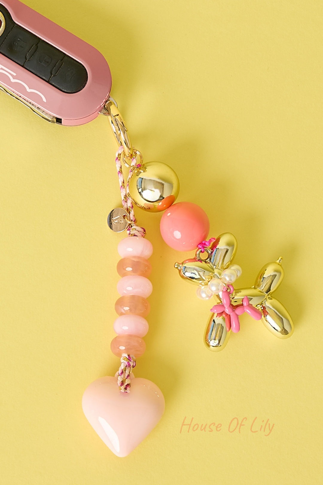 Light pink heart bag charm with peach and pink beads, gold-tone balloon dog charm attached, hanging next to pink car key fob on yellow background.