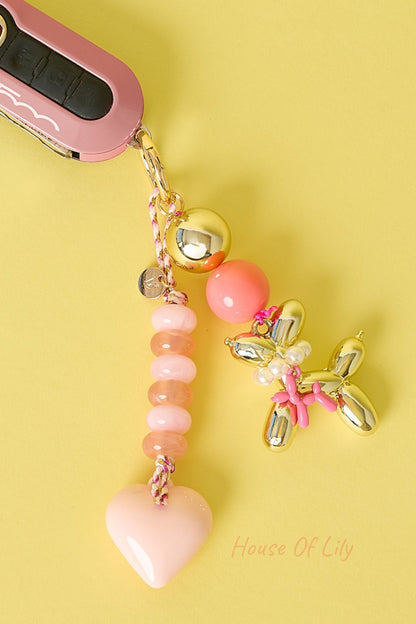 Light pink heart bag charm with peach and pink beads, gold-tone balloon dog charm attached, hanging next to pink car key fob on yellow background.