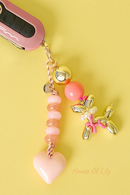 Light pink heart bag charm with peach and pink beads, gold-tone balloon dog charm attached, hanging next to pink car key fob on yellow background.