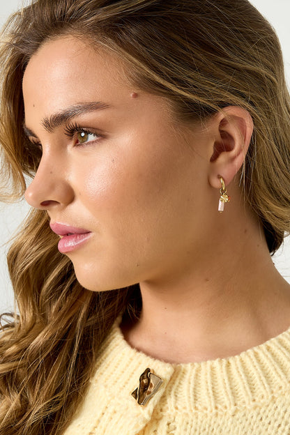 Woman with wavy hair wearing gold-tone hooped earrings with pink beads and white stone charms, wearing yellow knitted top