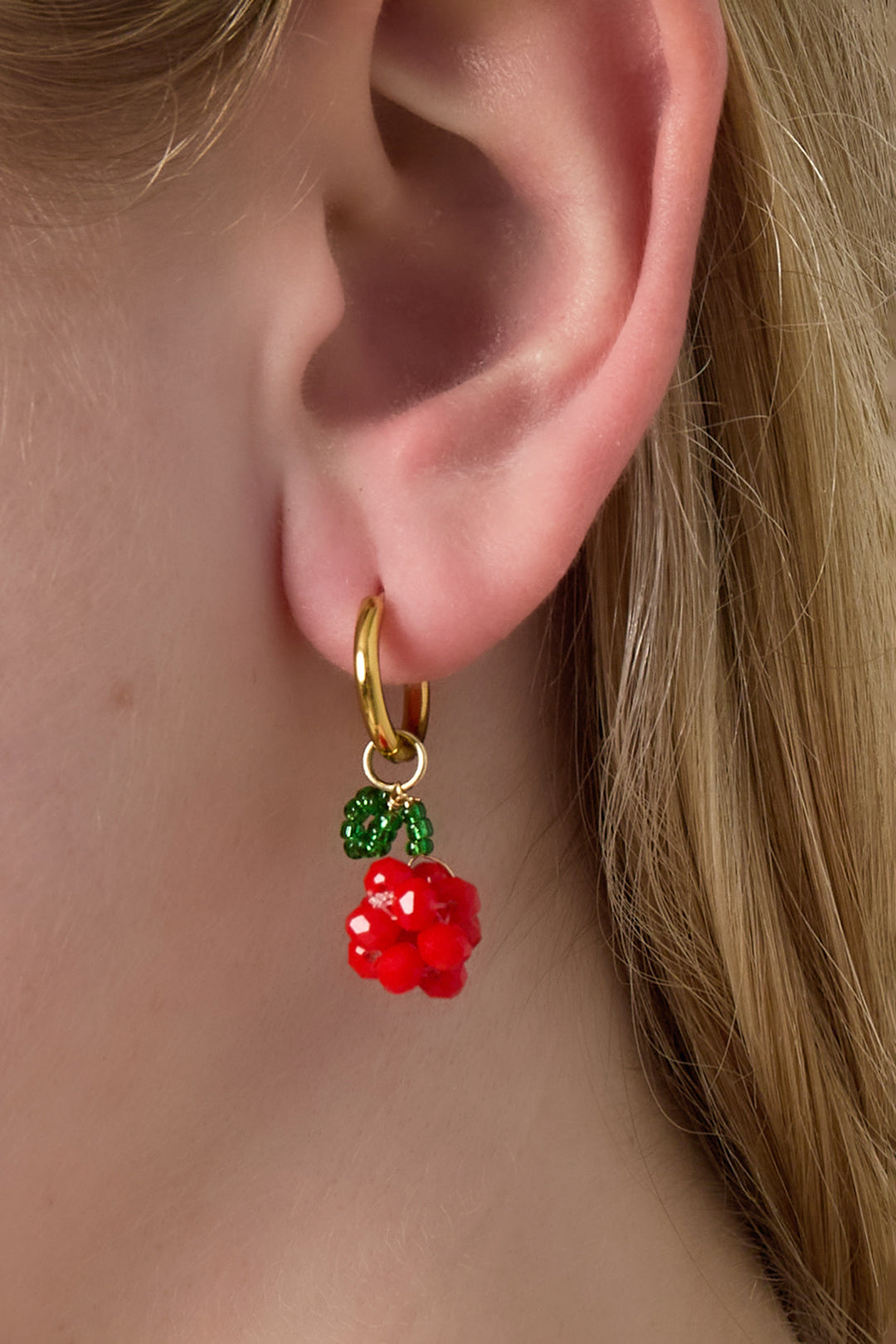Close-up of a woman's ear wearing a gold hoop earring with red beaded cherry cluster and green bead leaf charm