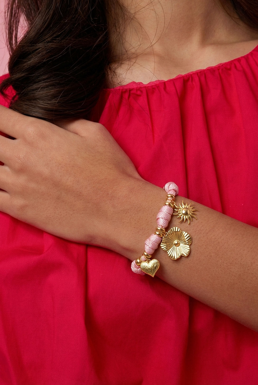 Model wearing pink rope charm bracelet with gold heart, flower, and sun charms paired with a red off-shoulder top
