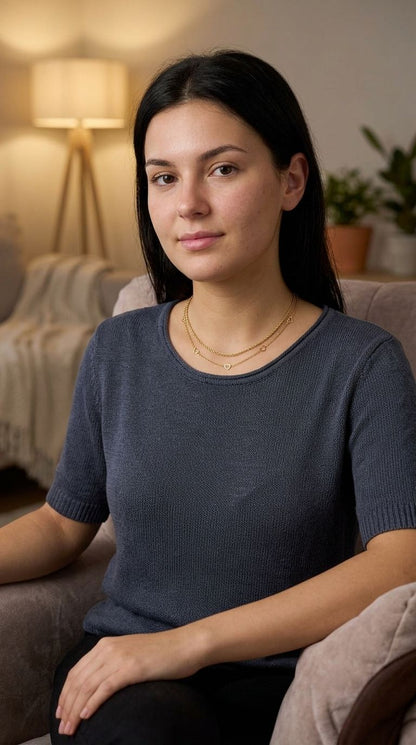 Model wearing a petite gold-plated heart chain necklace on her collarbone in a warm, softly lit indoor setting.