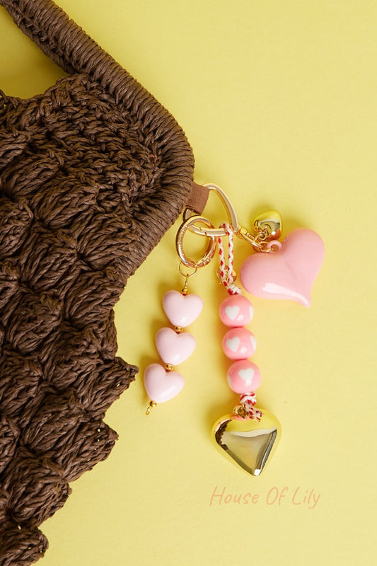 Brown woven bag with multiple heart-shaped bag charms in soft pink and gold tones against yellow background