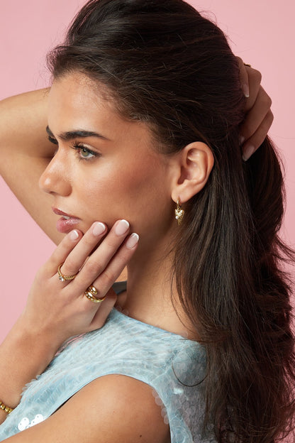 Model wearing gold heart-shaped hoop earrings with a small pearl accent, styled with matching gold rings.