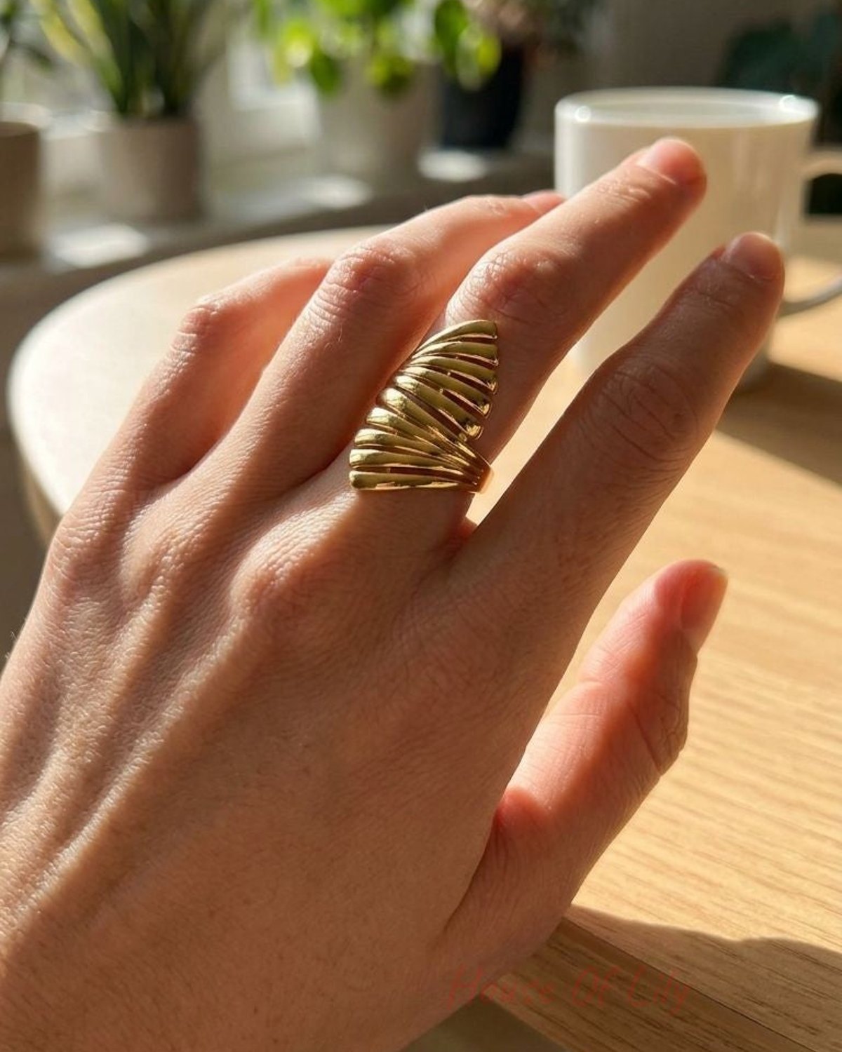 Worn on a slim finger, the Sculpted Gold Statement ring rests on a light wooden surface, showcasing its flowing wave design in natural sunlight.