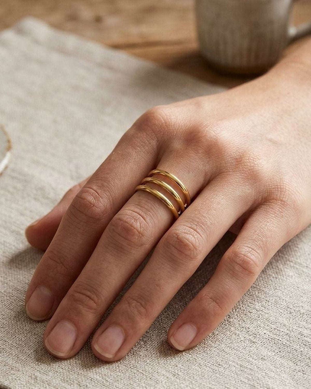 Model's hand wearing the 'Triple Layer Gold' ring with three sleek bands, set against a minimalist neutral-toned background with natural light, embodying everyday elegance.