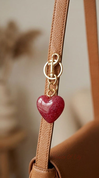 Deep red acrylic glitter heart bag charm with gold clasp hanging from a textured leather strap, showcasing shimmering surface and fine details.