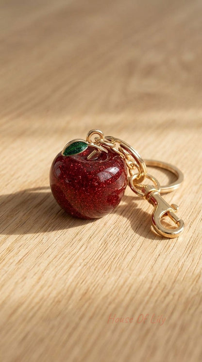 Red enamel apple bag charm with glitter accents and green leaf, attached to a leather strap with a gold clasp.