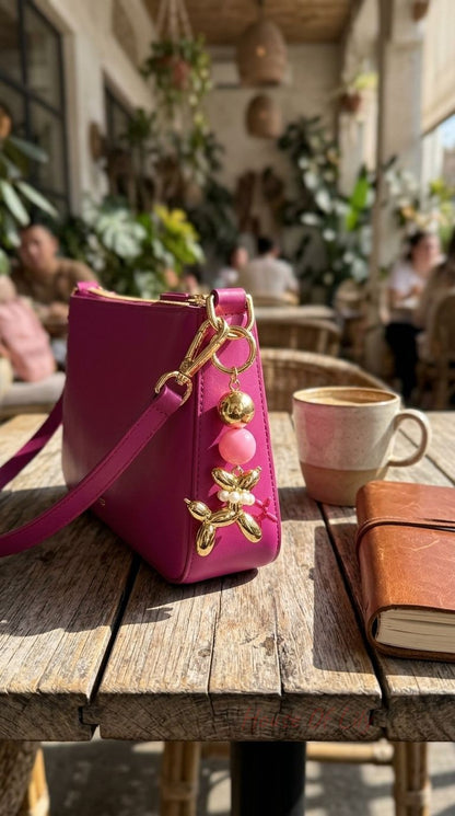 Gold balloon dog charm attached to a sleek bag placed on a rustic wooden table with natural morning light emphasizing its shiny finish and delicate pearl beads.
