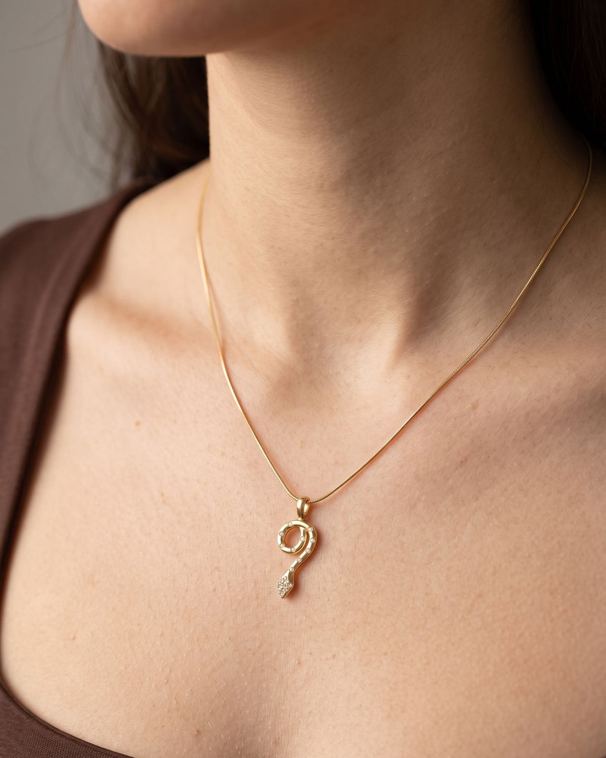 Woman wearing a minimalist gold snake pendant necklace with small rhinestones on a thin chain against a dark top.