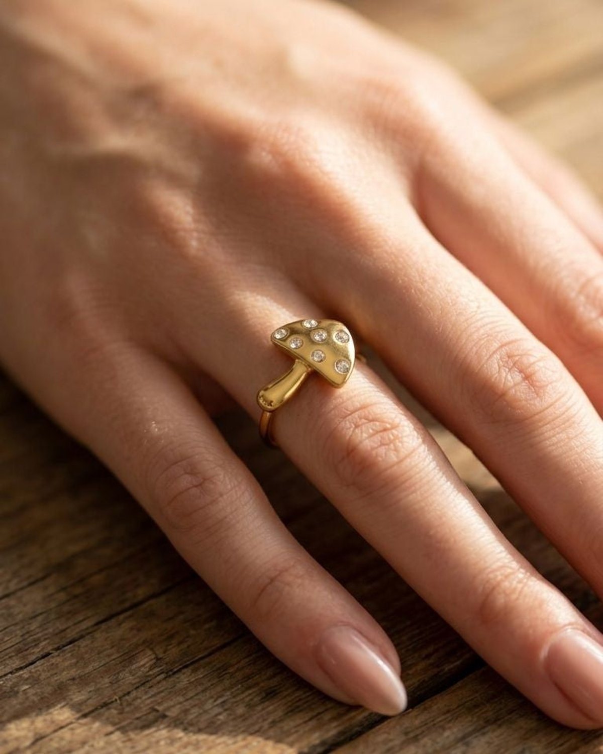 Full view of hand wearing Golden Mushroom Statement Ring on a rustic wooden surface, illuminated by warm, diffused sunlight showing whimsical mushroom shape.