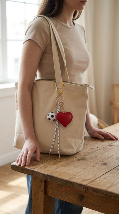Full-body view of the Heart & Soccer Bag Charm clipped onto a beige tote bag zipper, highlighting the gold hardware and red heart in a cozy room setting.