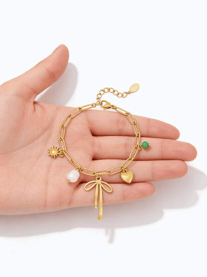 Gold chain bracelet with bow, heart, sun, pearl, and green bead charms held in a palm.