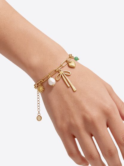 Gold chain bracelet on a wrist, featuring charms: a bow, pearl, heart, sun, and green bead.