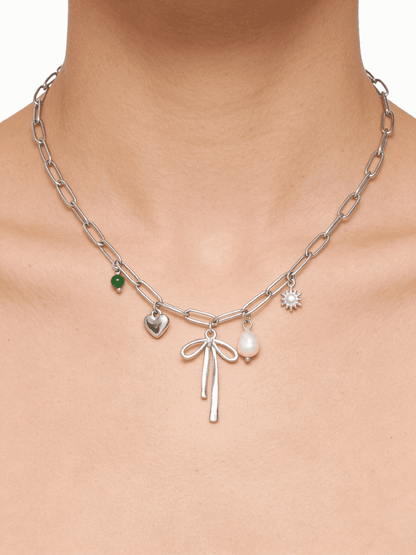 Silver link chain choker with a central bow charm, featuring a green bead, silver heart, pearl, and sun pendant.