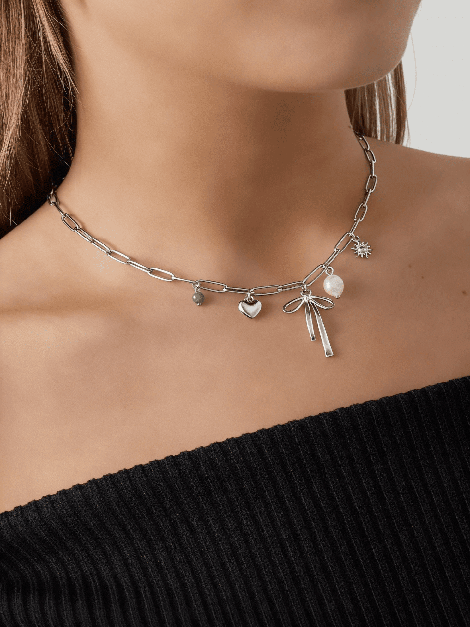 Silver chain choker featuring charms: a bow, heart, green bead, pearl, and sun, worn on a neck.