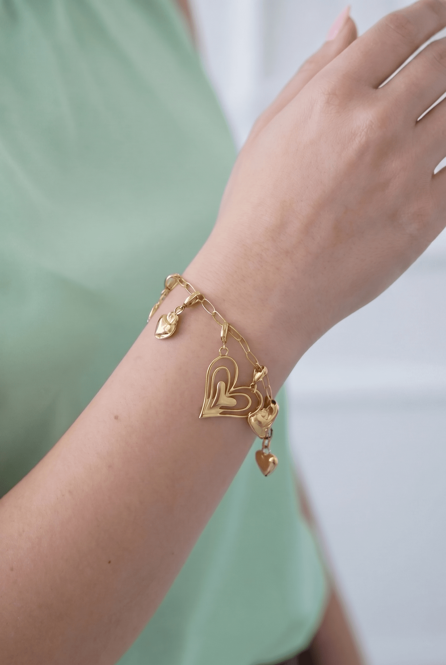 Gold charm bracelet on a wrist, featuring dangling heart charms of various shapes and sizes on a delicate link chain.