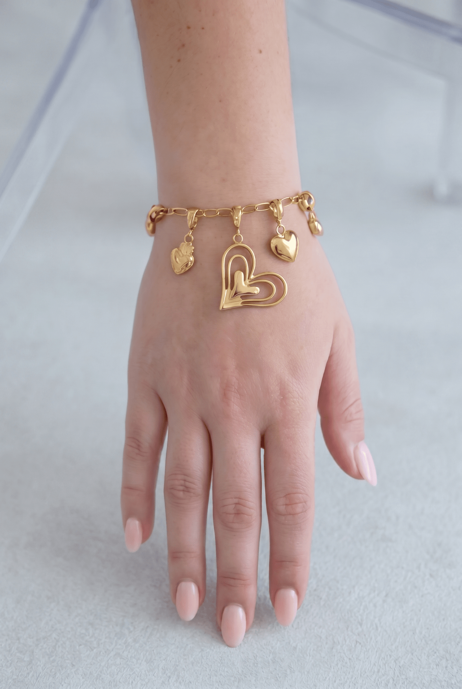 Gold chain link bracelet on a wrist, featuring a large open-heart charm and smaller solid gold heart dangles.
