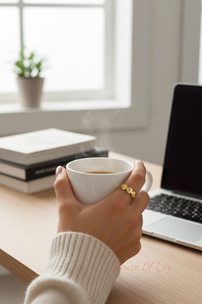 Gold heart ring featuring a continuous chain of small, polished gold hearts on a hand holding a steaming coffee cup.