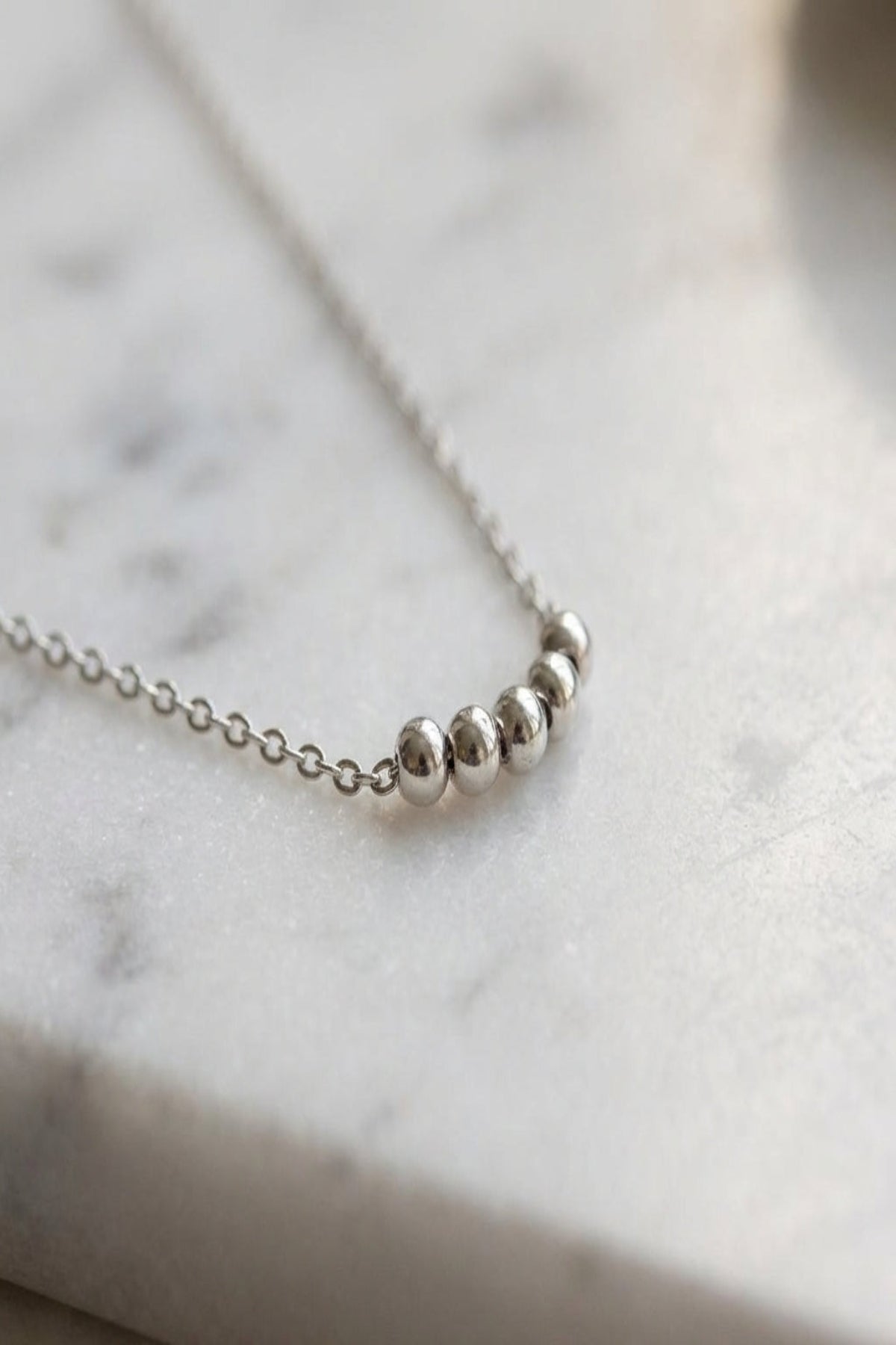 Close-up of a minimal ball chain necklace with tiny reflective discs on textured white marble, highlighting its delicate craftsmanship and mirror finish.