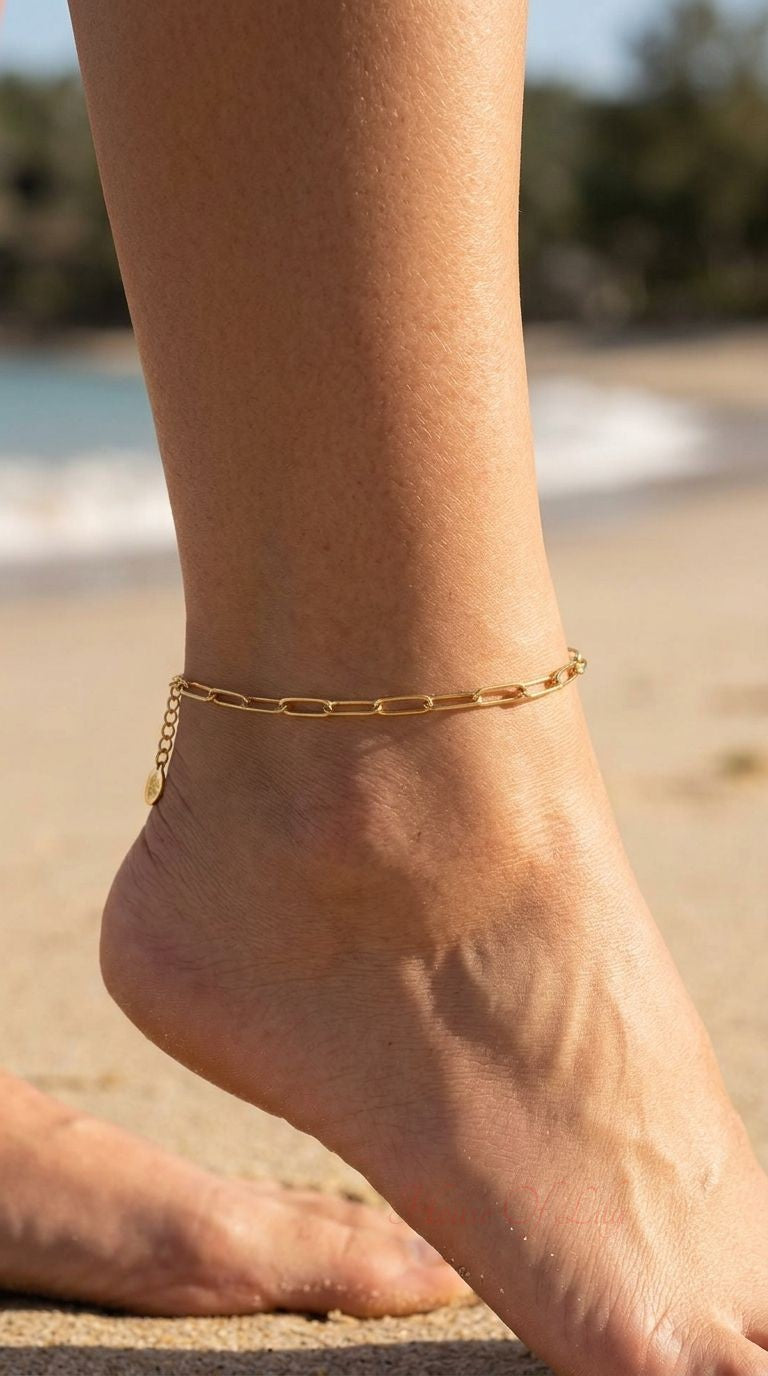 Close-up of a gold stainless steel chain link anklet on a sun-kissed ankle in an outdoor setting with beach and foliage background.