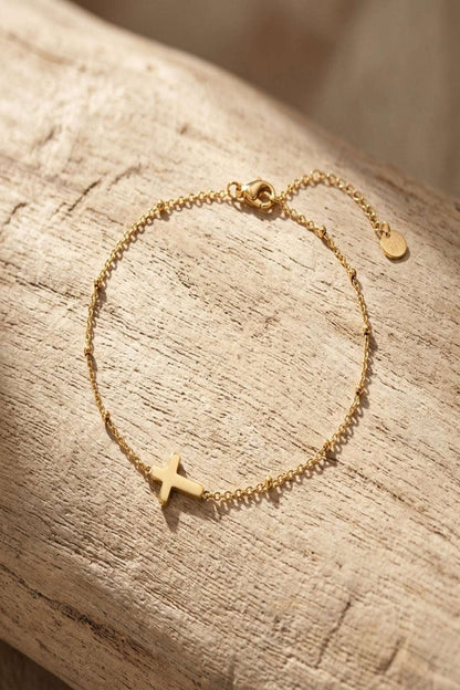 Close-up of a minimalist gold cross stainless steel bracelet on a textured wooden surface, showcasing its delicate design and intricate charm.