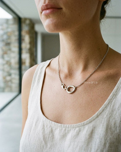 Minimalist silver oval clasp chain necklace worn around the neck against a soft-focus architectural background.