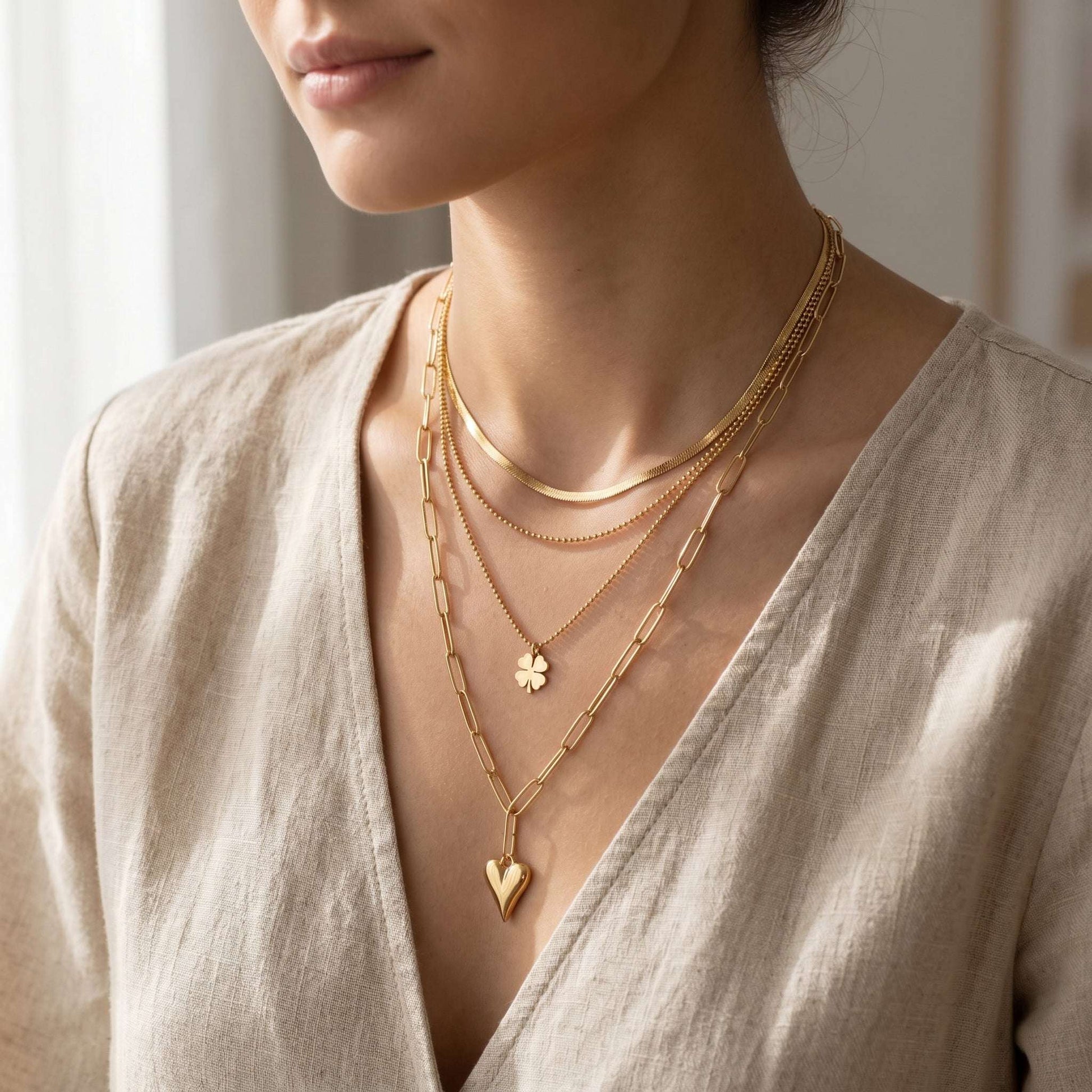 Close-up of a delicate three-layer stainless steel necklace with heart and clover pendants against a textured fabric background.