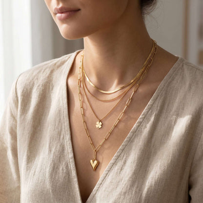 Close-up of a delicate three-layer stainless steel necklace with heart and clover pendants against a textured fabric background.