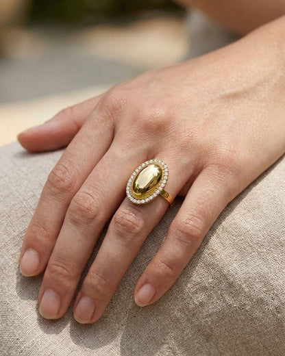 A hand wearing the Golden Oval Statement ring on the middle finger, showing its sparkle and delicate design against a rustic fabric background.