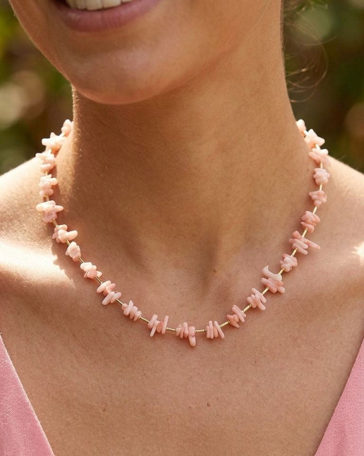 Close-up of pink coral chip necklace with gold accents worn by a smiling woman, highlighting delicate coral and sunlit outdoor background.