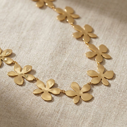 Detailed close-up of a gold flower necklace with micro-texture, draped on beige fabric, showcasing delicate craftsmanship and subtle shine.