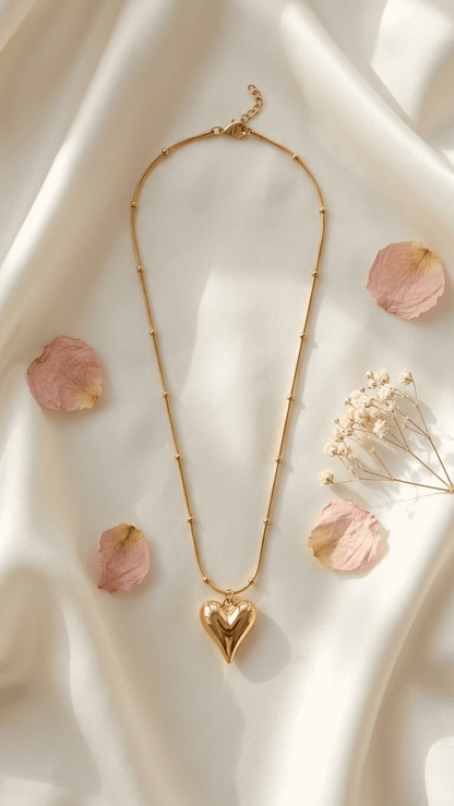 Gold-tone necklace with a puffed heart pendant and bead-detailed chain on a draped cream silk fabric with flower petals.