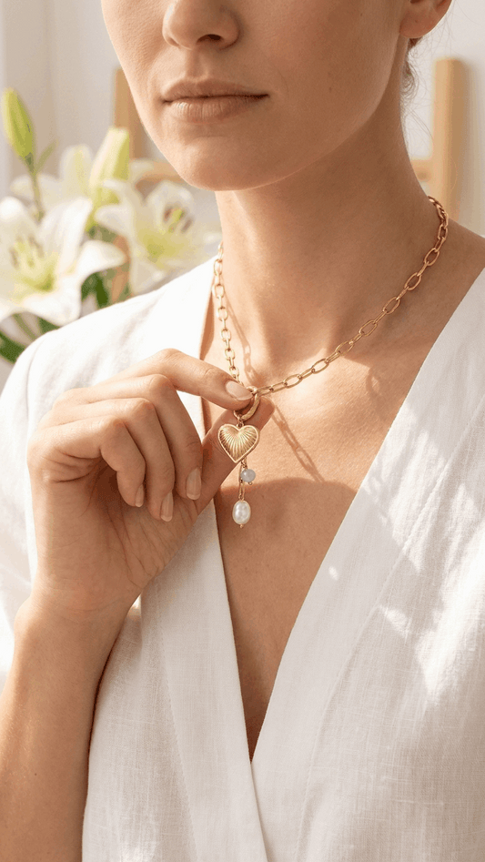 Gold chain necklace with a ribbed heart pendant, small blue bead, and a white pearl charm worn by a woman.