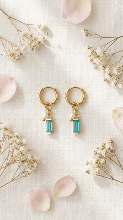 Dainty gold hoop earrings with a dangling rectangular teal gemstone and a small white pearl, on a floral background.