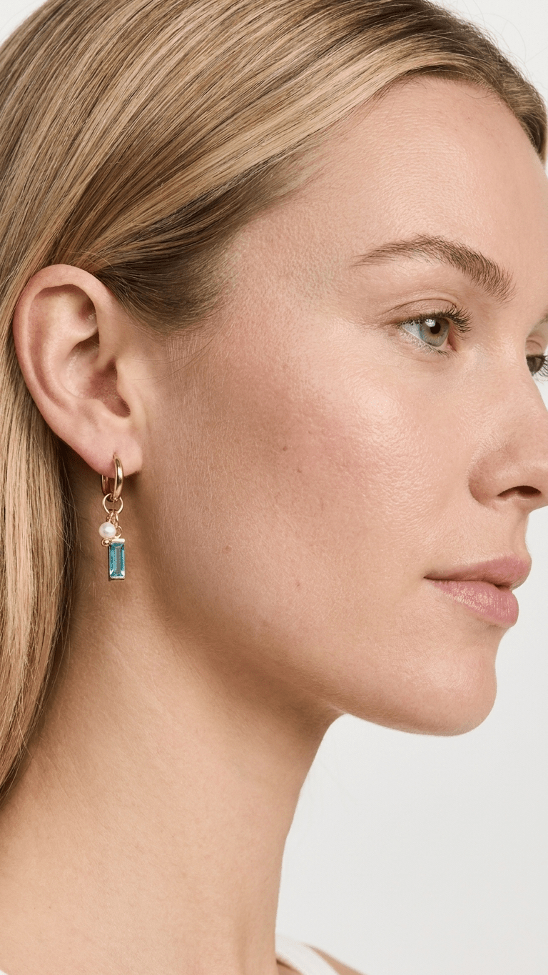 A model wears gold hoop earrings with dangling charms of a small white pearl and a rectangular blue crystal.