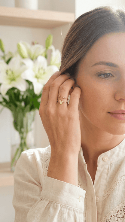 Dainty gold ring with a heart charm and dual-band open design, worn by a woman next to white lilies.