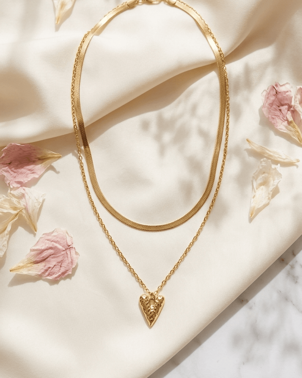 Gold layered necklace with a herringbone chain and a chain holding a textured heart pendant on a white fabric.