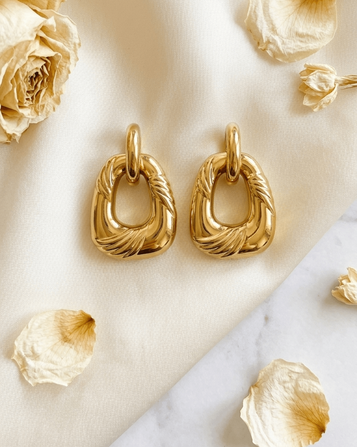 Gold chunky geometric hoop earrings with ridged texture, resting on cream silk with dried rose petals.