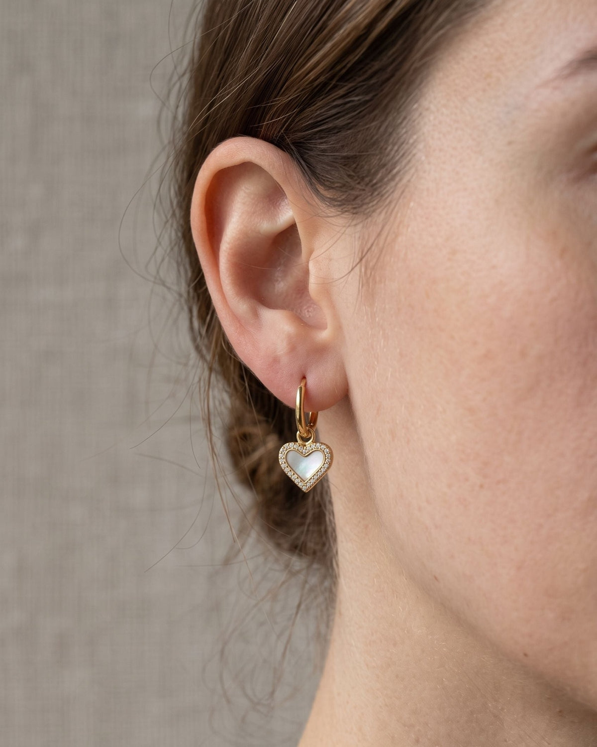 Delicate heart drop earrings featuring sea shell inlay and zircon accents worn on an earlobe.