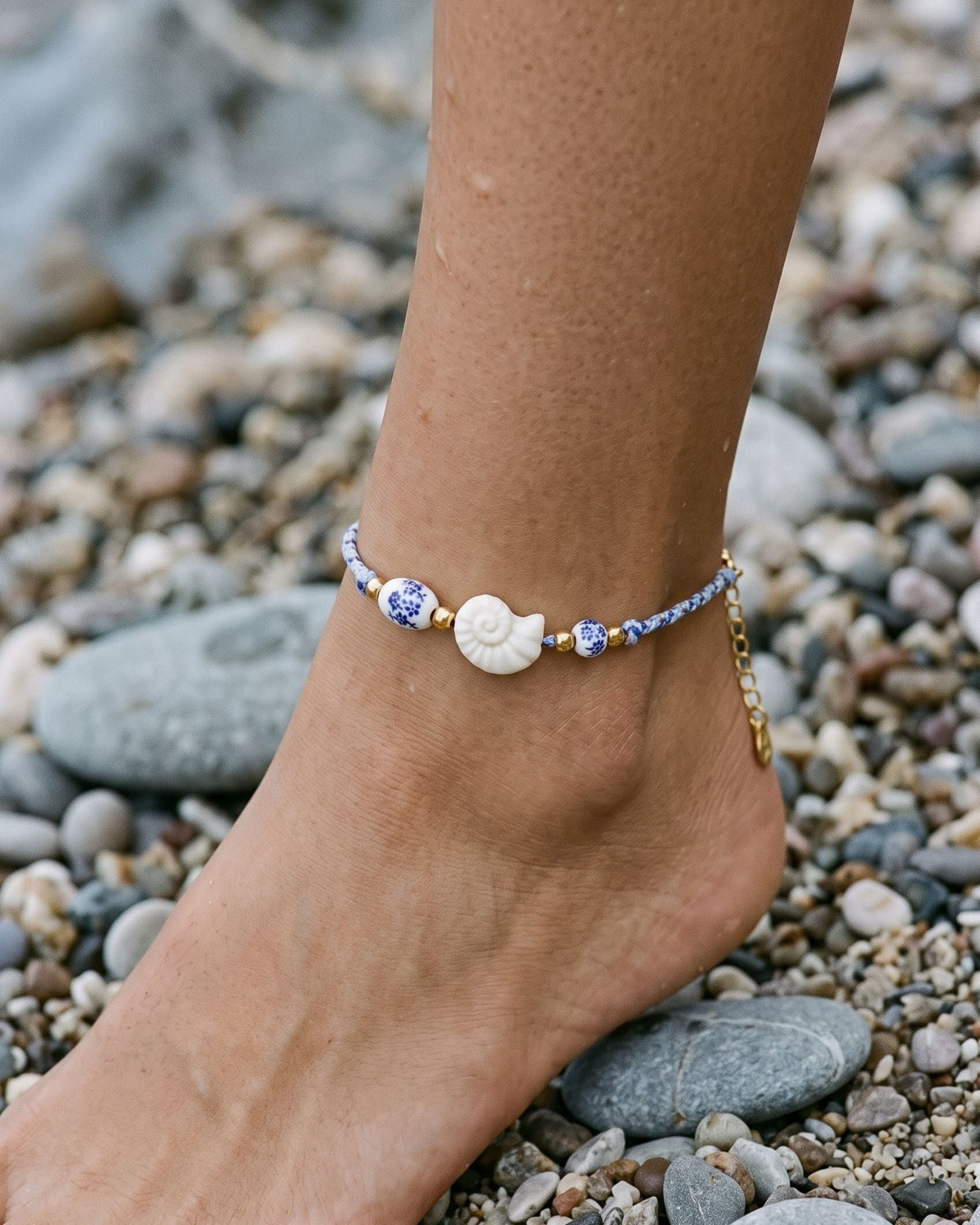 Shell and ceramic bead anklet with gold-toned spacers on braided rope worn on an ankle over gray beach pebbles.