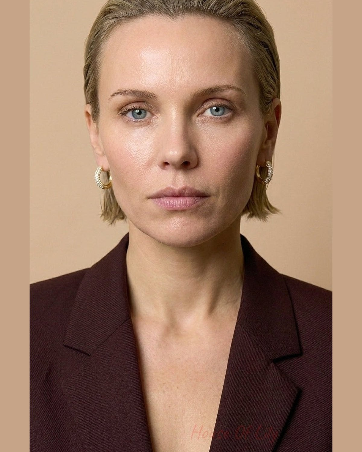 A woman with slicked-back blonde hair and a maroon blazer wears pavé-set gold hoop earrings.