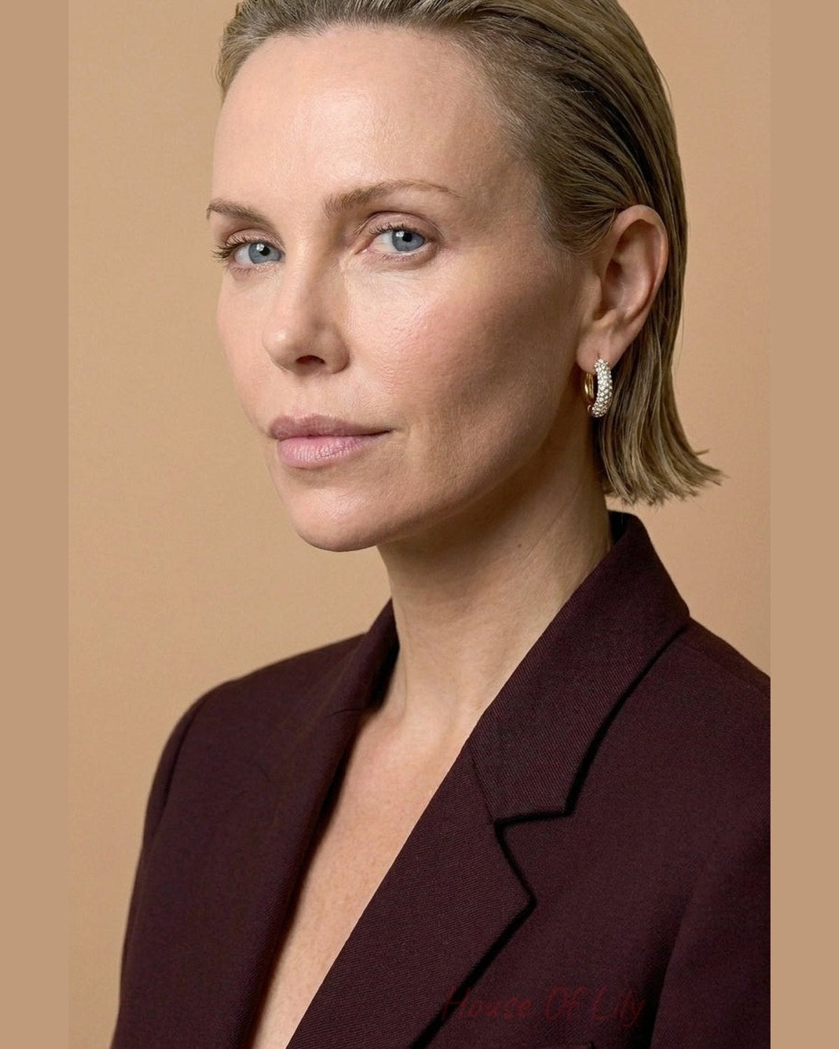 A person with slicked-back blonde hair and a maroon blazer wears gold hoop earrings.