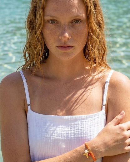 Woman in white top wearing a braided orange cord bracelet with gold charms, pearl, and red bead.