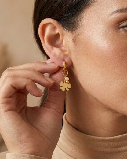 A close-up of a woman wearing a delicate gold hoop earring with a dangling gold four-leaf clover charm, paired with a beige turtleneck sweater.