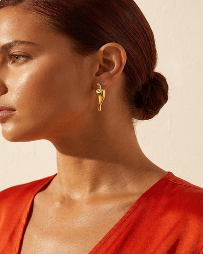 A close-up side profile of a woman with dark hair in a bun wearing a delicate gold drape earring, paired with a vibrant orange-red top against a warm beige background.