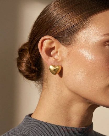 A close-up side profile of a woman wearing a small, polished gold heart-shaped stud earring. Her hair is styled in a low bun, and she is wearing a dark gray ribbed turtleneck sweater against a warm, softly lit beige background.